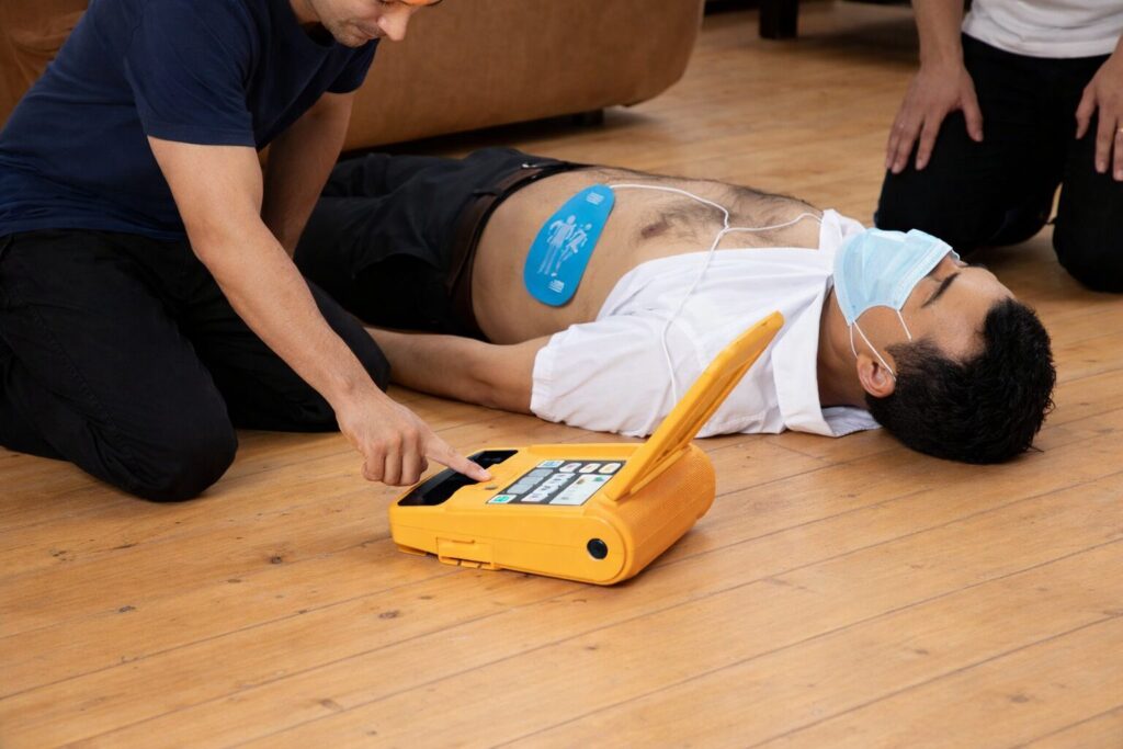Two trained responders applying AED electrode pads and performing CPR on a man in an office setting using the Amoul i5 defibrillator.